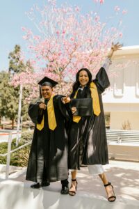 Two graduates celebrating with diplomas, wearing black gowns and yellow stoles outdoors.